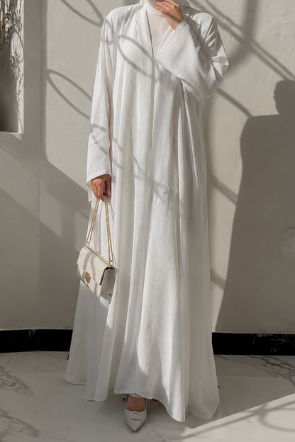 Woman in a white abaya standing in a softly lit room with shadows on the wall.