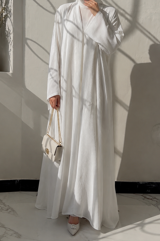 Woman in a white abaya standing in a softly lit room with shadows on the wall.